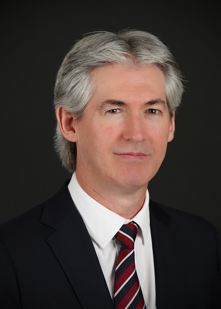A Caucasian man with grey hair wearing a black suit, white shirt, red, white and navy striped tie smiles softly at the camera