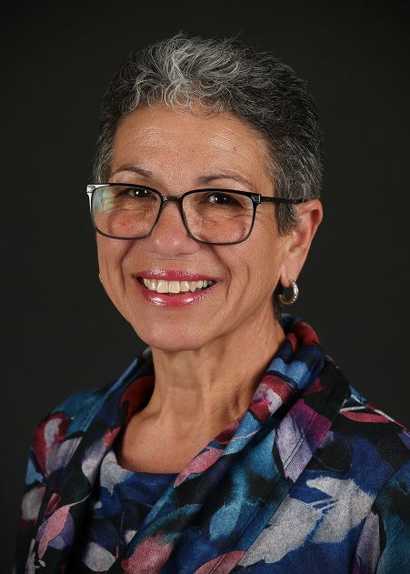A Caucasian woman with short grey hair wear glasses and a blue, pink and white patterned top smiles at the camera.